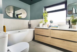 Bathroom with grey floor and wall tiles, blue painted wall and ceiling, wood effect vanity unit, white freestanding bath and oval mirror