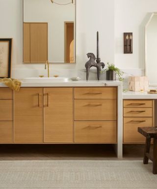 A beige bathroom with a rectangular wall mirror, a wooden vanity with plants and candles on it, and a white woven rug underneath it