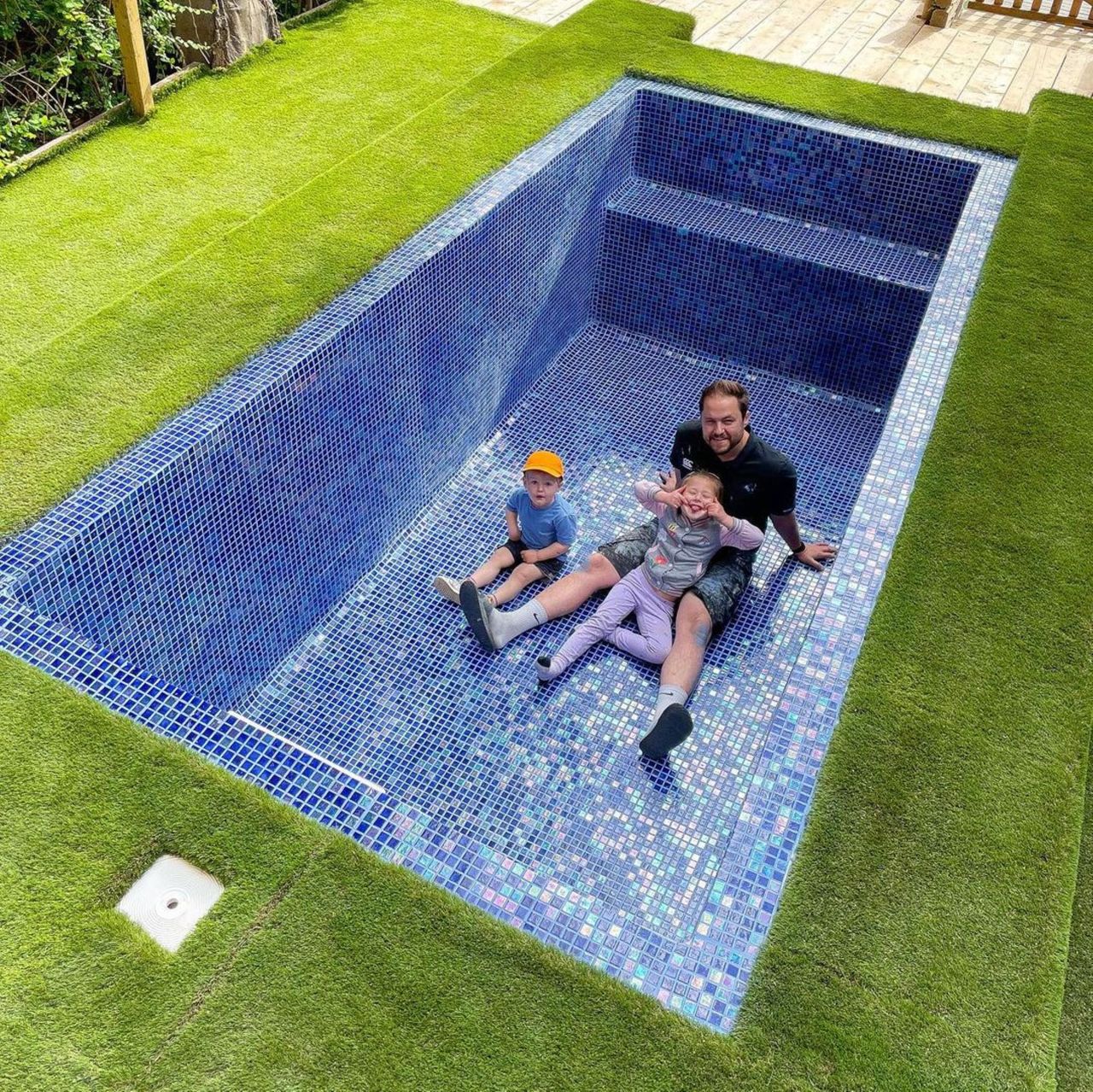 The DIY pool with Alex with his children in the pool which isn't filled surrounded by Astroturf