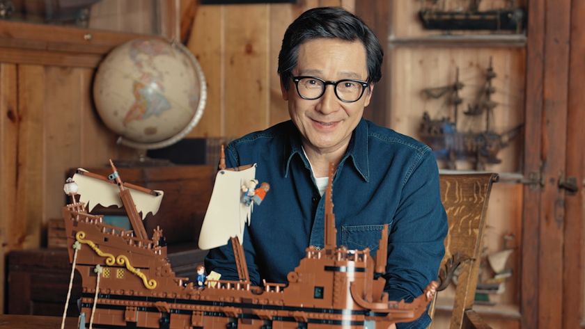 Ke Huy Quan smiles while sitting behind a Lego Goonies set, in a nautically-themed wood-panelled room