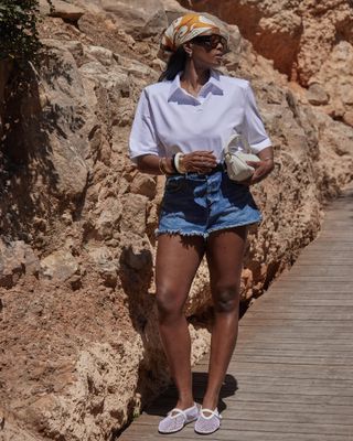 British fashion influencer Marilyn Nwawulor-Kazemaks poses while on vacation in Ibiza, Spain wearing a printed silk head scarf, chunky aviator sunglasses, a white polo t-shirt, denim cutoff shorts, cream mini shoulder bag, and white Alaia mesh flats