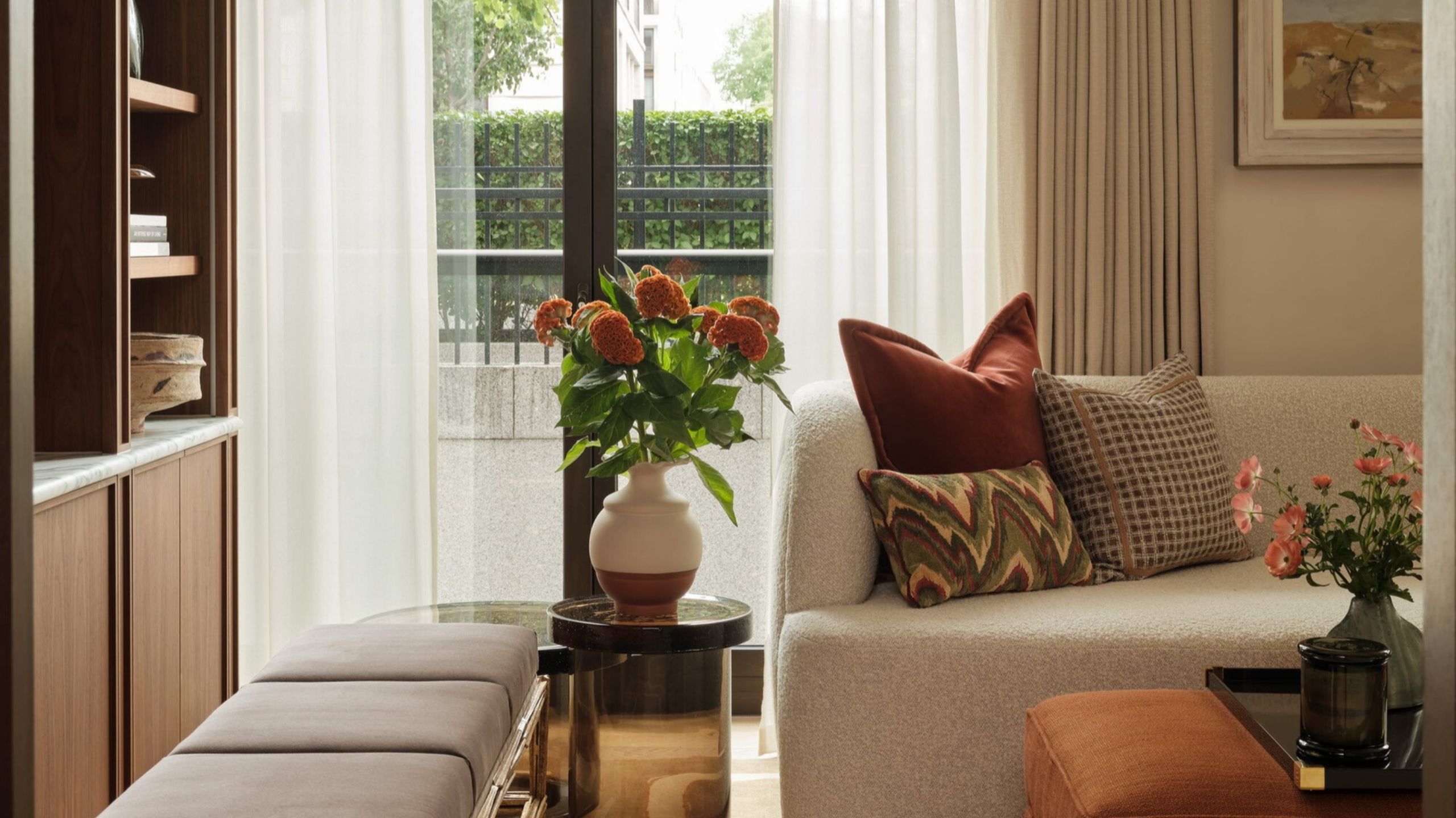 a cozy living room with doors leading to outside with sheer curtains, a built-in bookcase, a bench seat next to a side table with a vase of flowers, beige sofa with colored cushions, and ottoman