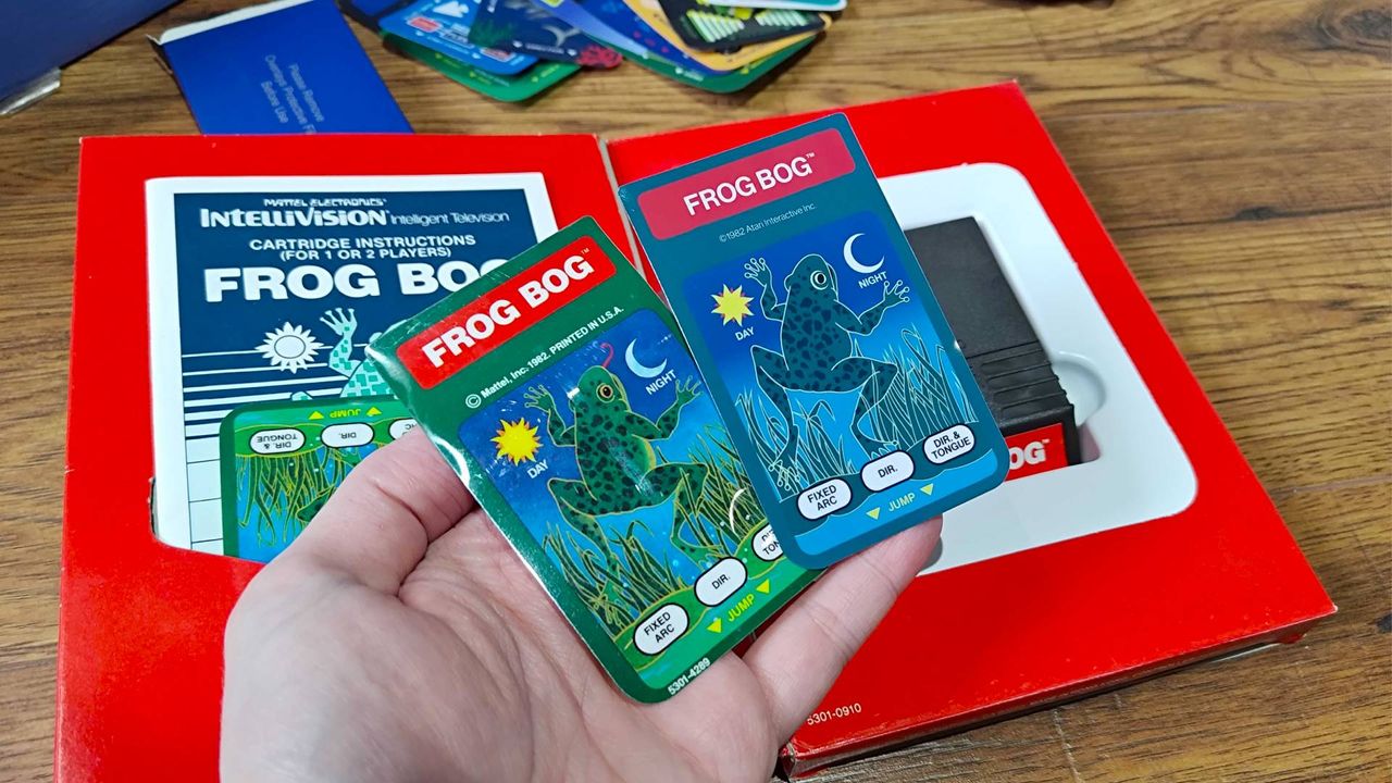 Hand holding Frog Bog overlay for both Intellivision Sprint and original cartridge with box in backdrop.
