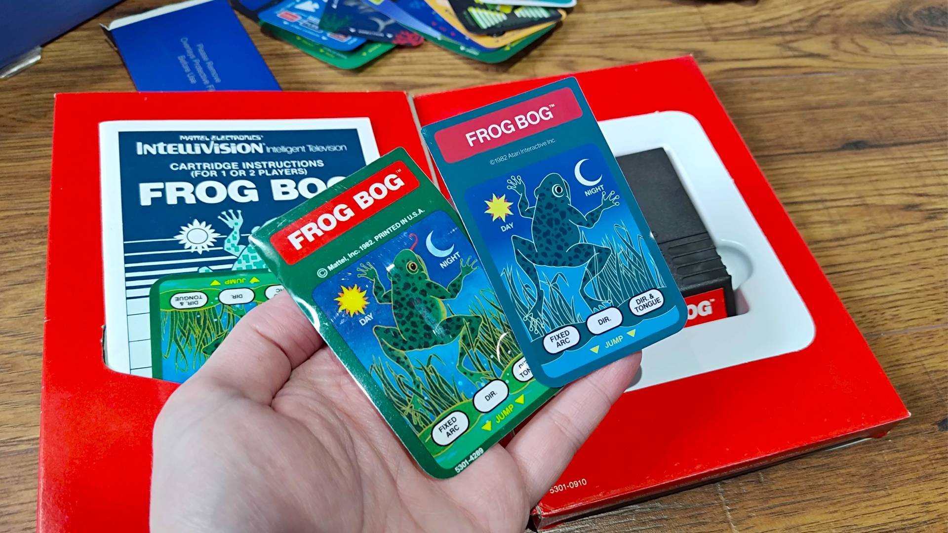 Hand holding Frog Bog overlay for both Intellivision Sprint and original cartridge with box in backdrop. 