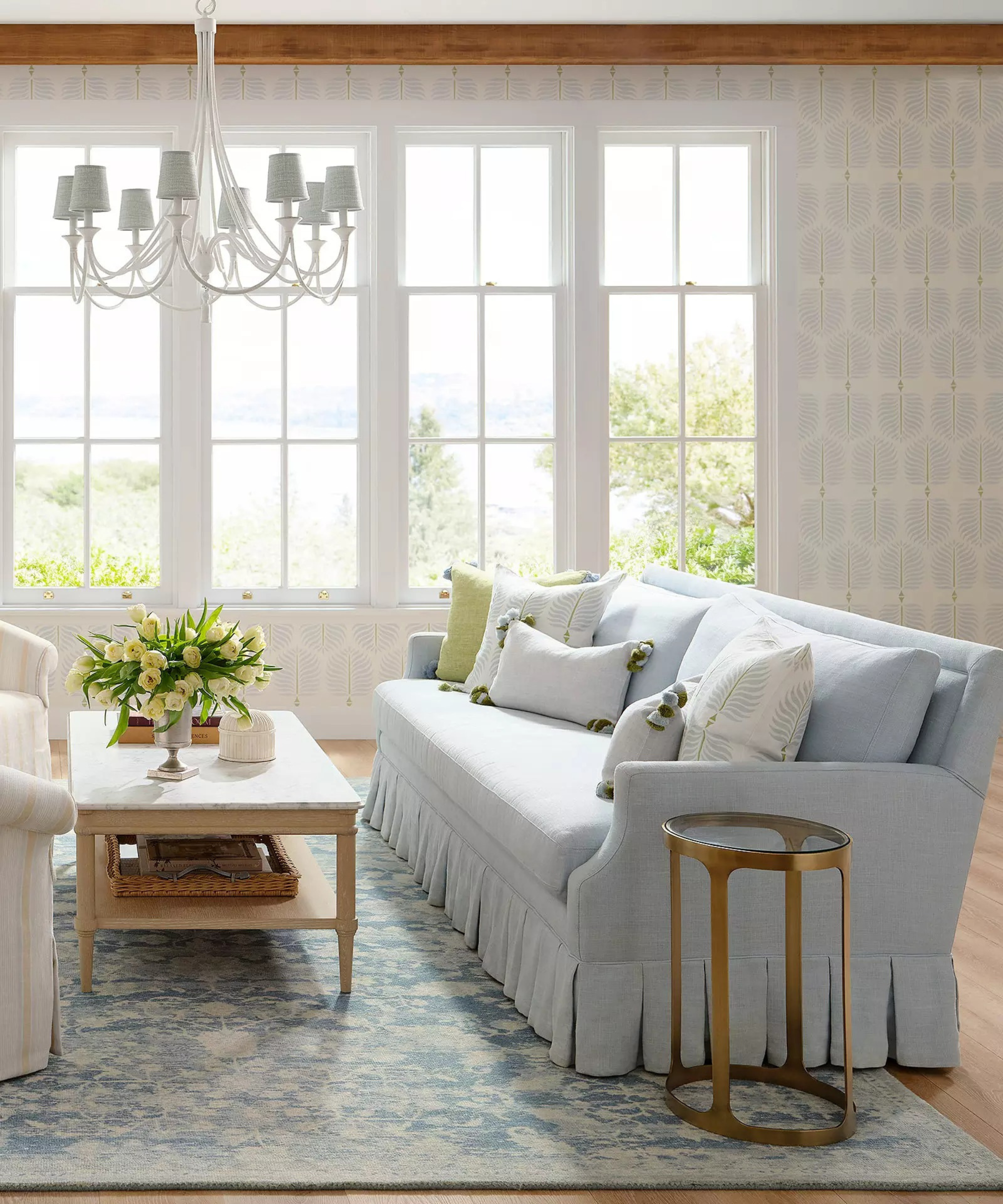 a coastal living room with a large blue couch with pillows on top, a coffee table with a vase of flowers on it, a brass finished side table, a chandelier hanging overhead, and windows in the background