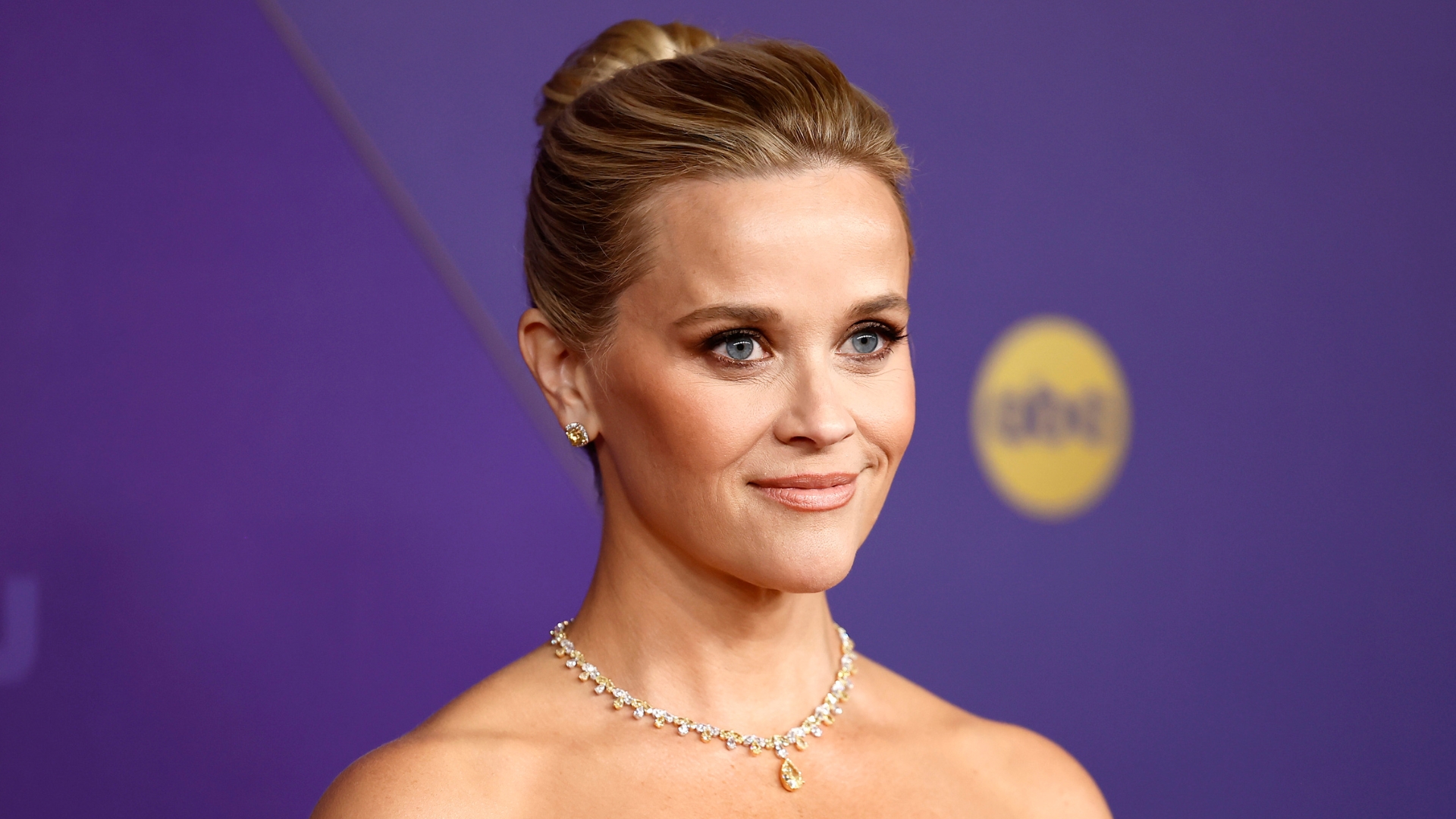 Image of Reese Witherspoon smiling with her blonde hair in a bun and wearing a crystal necklace