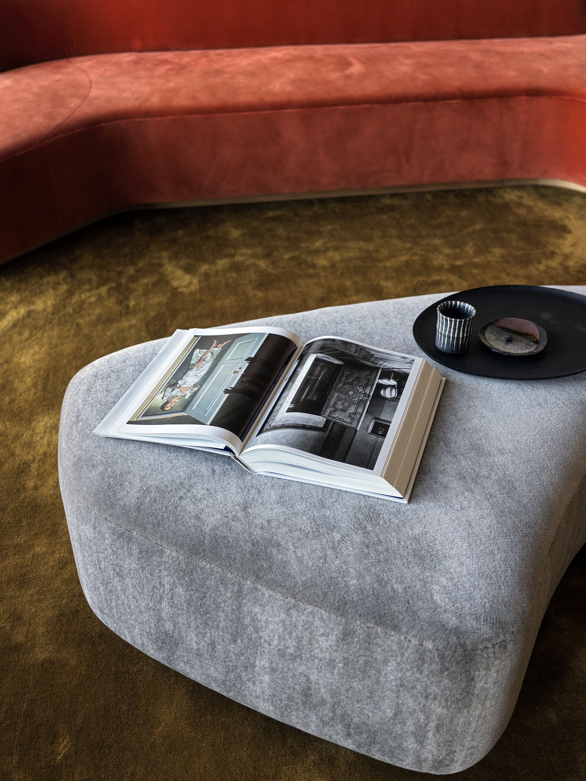 A gray ottoman has a photography book open on top of it next to a black tray in front of a res sofa on an ochre carpet