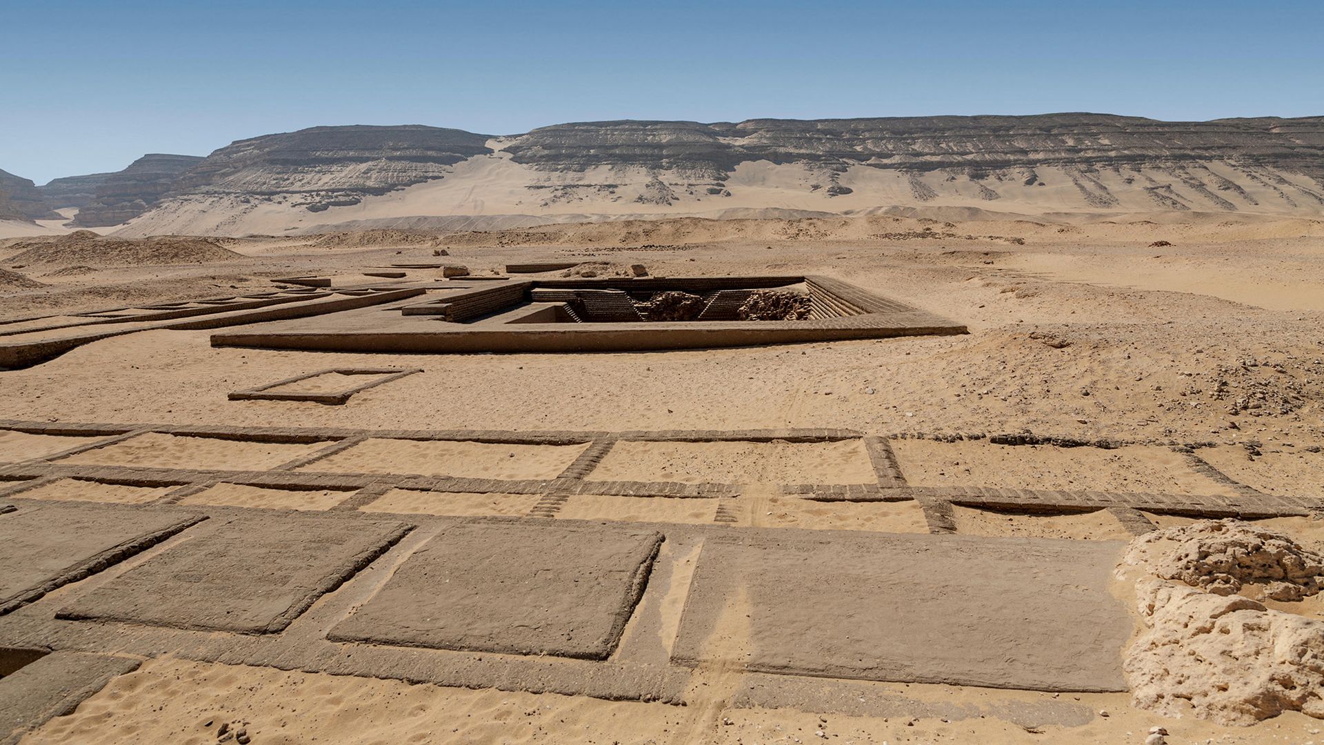 A series of square areas are seen in the desert, with a larger tomb in the background