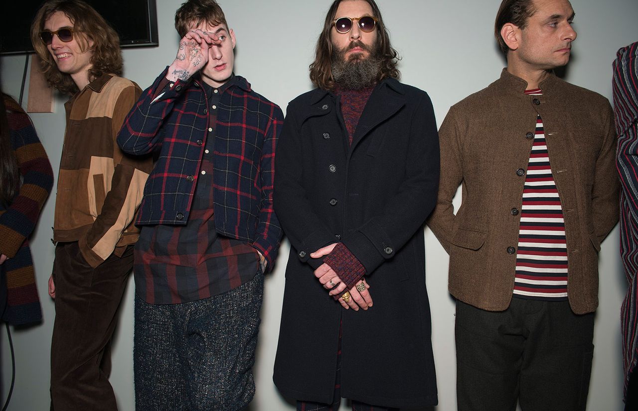 Four male models wearing looks from Oliver Spencer&#039;s collection. One model is wearing sunglasses, brown trousers and a jacket with sections in different shades of brown. Another model is wearing a blue and red plaid jacket and red, grey and black check style shirt. Next to him is a model wearing a red jumper and black coat. And the fourth model is wearing a brown jacket, dark trousers and red, white and black horizontally striped top