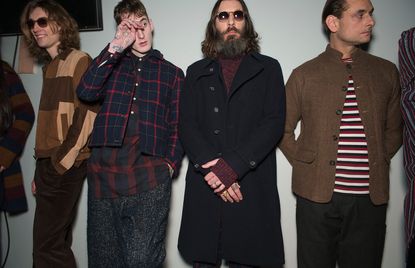 Four male models wearing looks from Oliver Spencer's collection. One model is wearing sunglasses, brown trousers and a jacket with sections in different shades of brown. Another model is wearing a blue and red plaid jacket and red, grey and black check style shirt. Next to him is a model wearing a red jumper and black coat. And the fourth model is wearing a brown jacket, dark trousers and red, white and black horizontally striped top