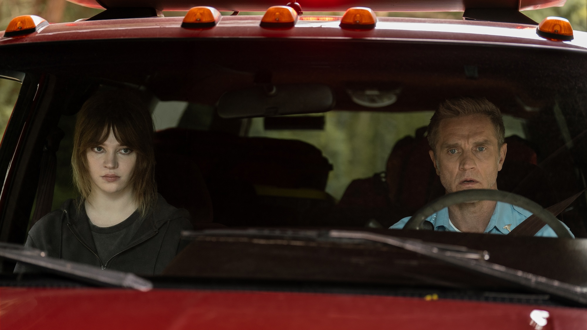 Sophie Thatcher's Nat looking unhappy while inside a red truck driven by Devon Sawa's Howard wearing blue shirt in Yellowjackets Season 4
