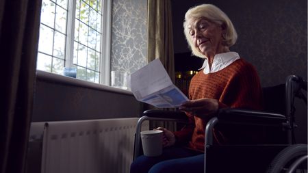 An older woman sits in a wheelchair by a window and looks at paperwork.