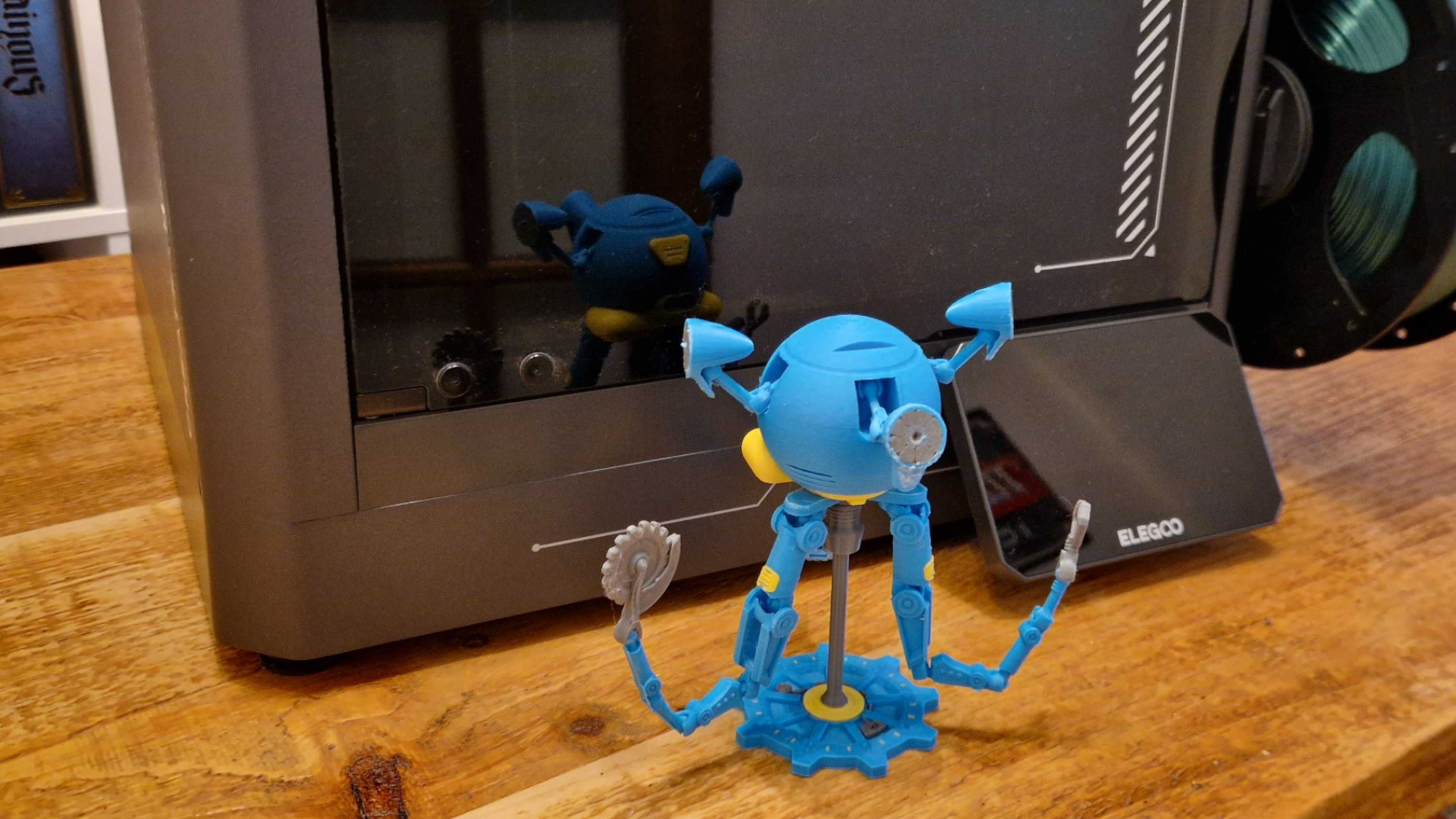 A blue and yellow Mr Handy model on a wooden table, in front of the Elegoo Centauri Carbon 2 Combo