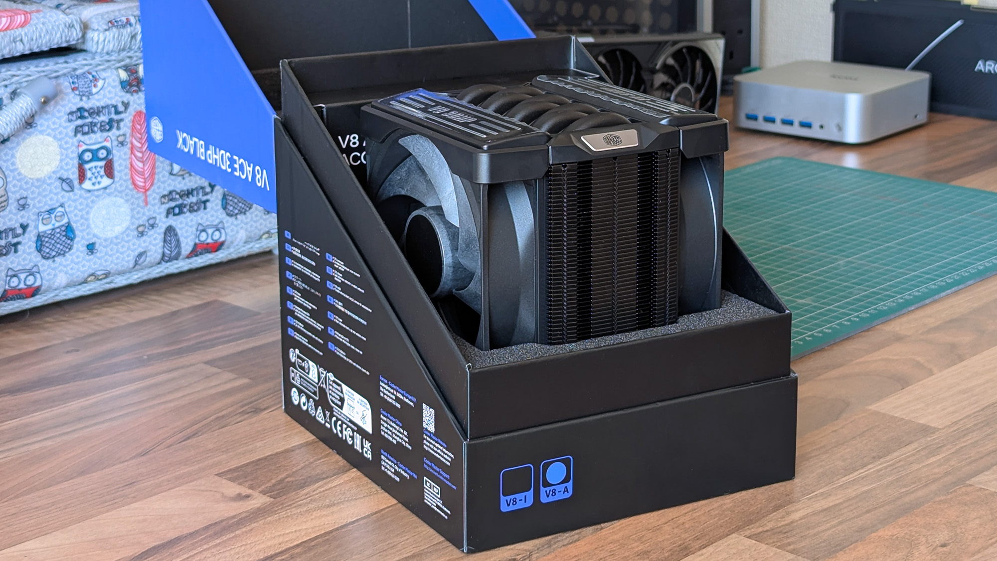 The Cooler Master V8 ACE 3DHP Black CPU cooler rests inside its open box, revealing its dual&amp;#8209;fan design and dense fin stack. Surrounding PC parts and tools sit on a desk with a green cutting mat.
