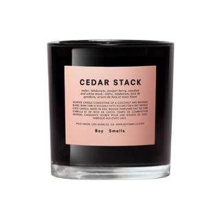 A black candle vessel with a peachy pink label on the front of it that says 'Cedar stack'