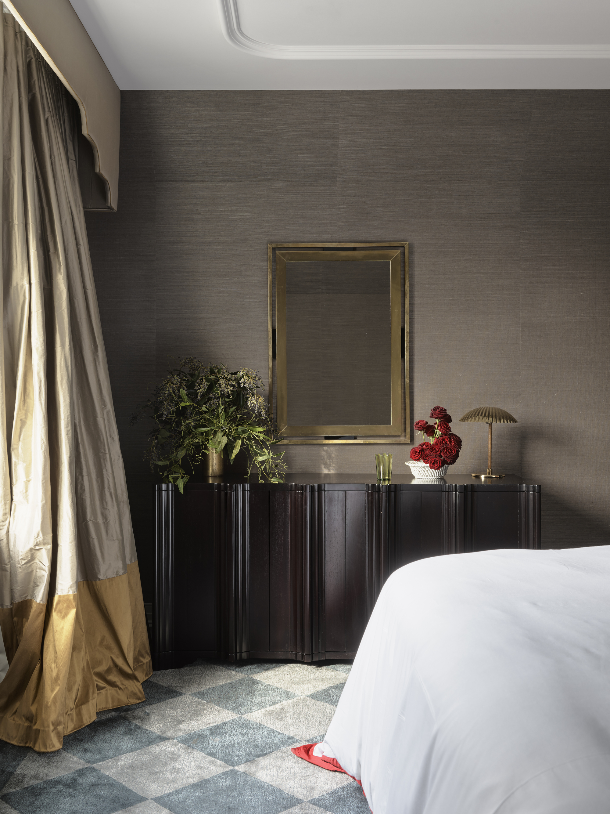 modern bedroom with brown grasscloth wallpaper, white ceiling, checkerboard rug, dark timber console styled with vase of flowers, lamp, and glass of water, with mirror hanging above, white bedcover, with pelmet over heavy two-tone curtains