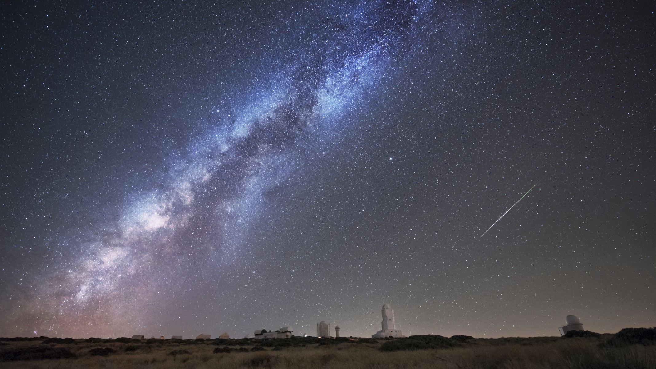 night sky landscape image with milky way stretching across the sky and a meteor streaking in the center right