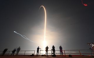 the silhouettes of several people can be seen as a rocket launch creates a streak of light in the background