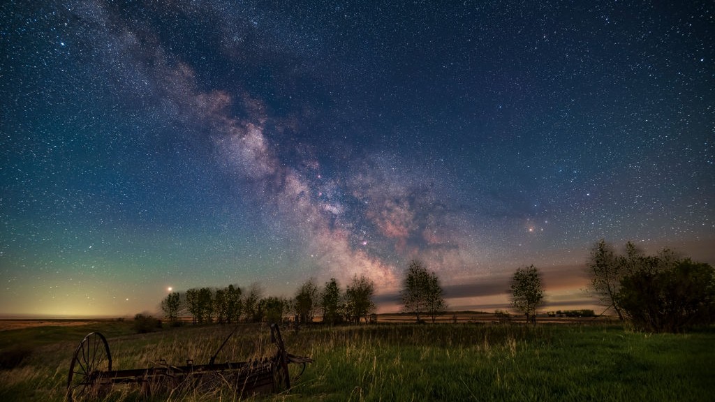 night scene showing milky way stretching across thy sky. trees are silhouetted below, there is a bright orange star on the right - Antares and two bright points of light on the left - Jupiter and Saturn.