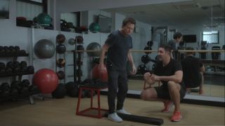 Michael J. Fox standing in a gym with his trainer squatting beside him.