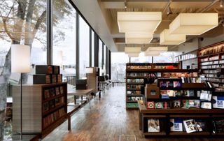 Interior view of Tsutaya Books featuring wood flooring, floor-to-ceiling windows, cuboid style ceiling lights, floor lamps and dark wood display and shelving units filled with items