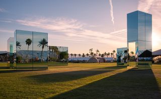 Mirrored cubes and rectangular objects on green grass with palm trees behind them.