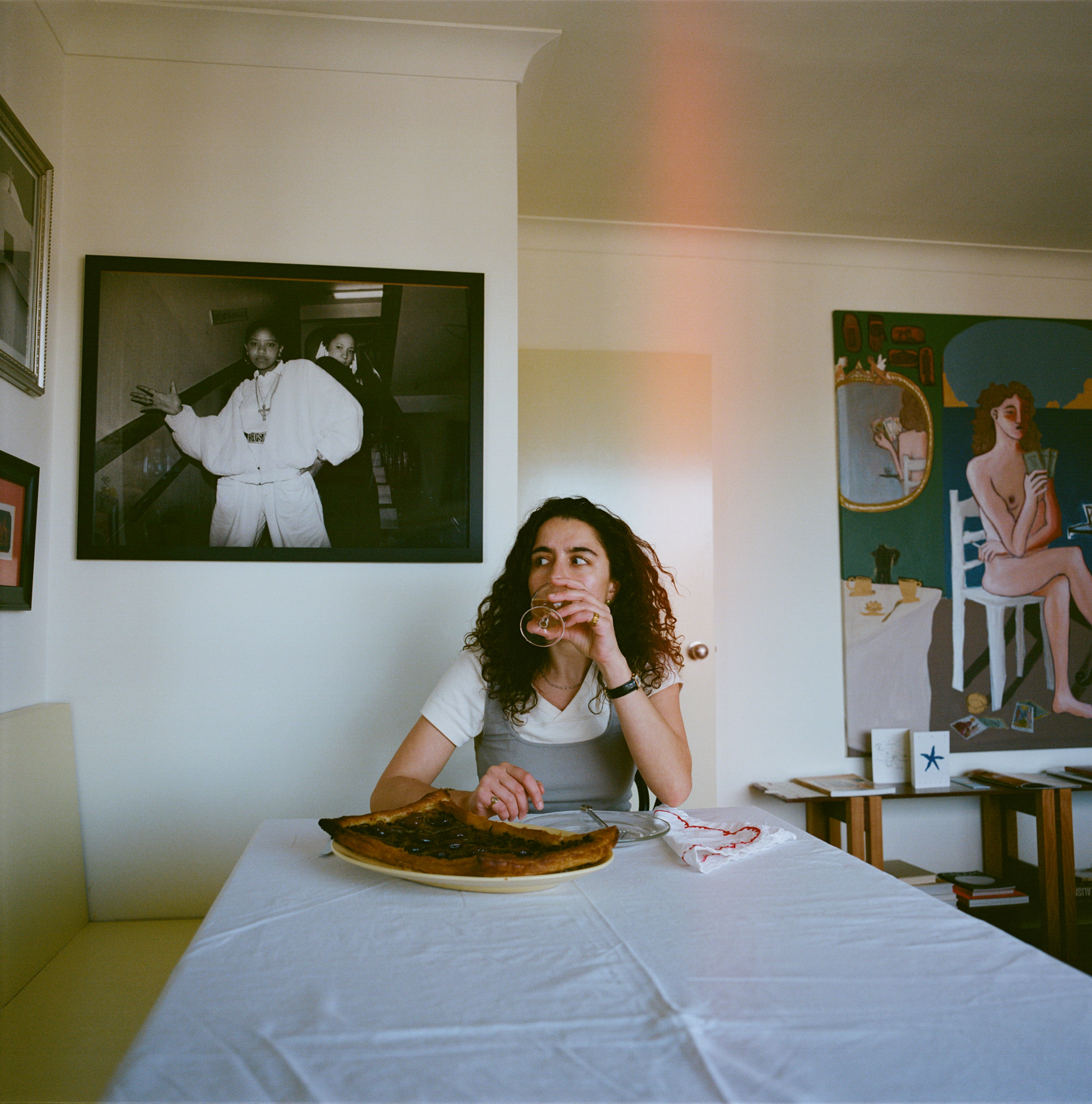 A young curly haired woman with a white and gray top sits at the dining table of her flat surrounded by art.