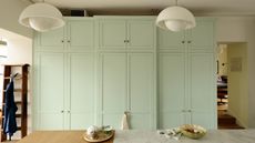 Six tall pastel green kitchen cabinets with a few inches gap at the top. There is a marble and wooden kitchen island in foreground with retro while ceiling lamps and a wooden ladder in view