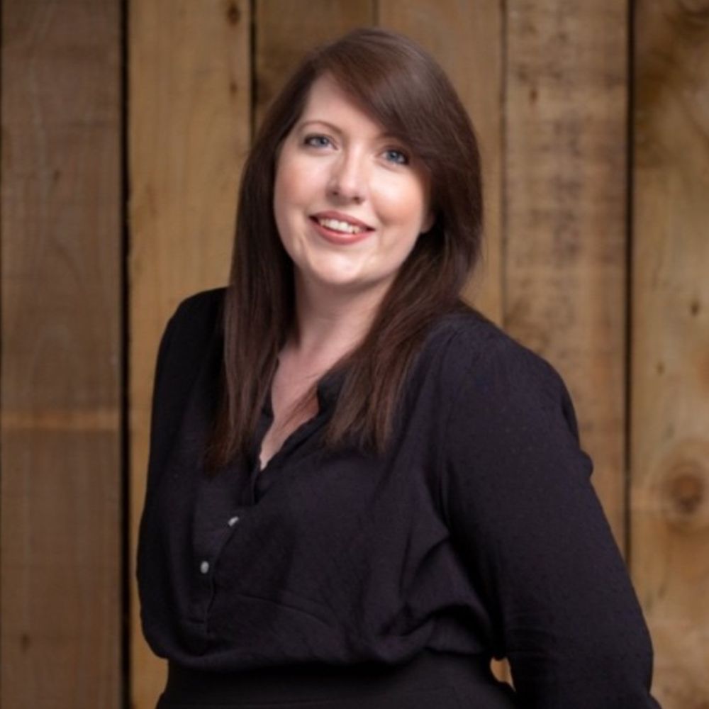 A head-and-shoulders portrait of a smiling woman with straight, shoulder-length brown hair parted to one side. She wears a loose black blouse. The background is made of vertical wooden planks, softly lit, giving the image a warm, natural feel.