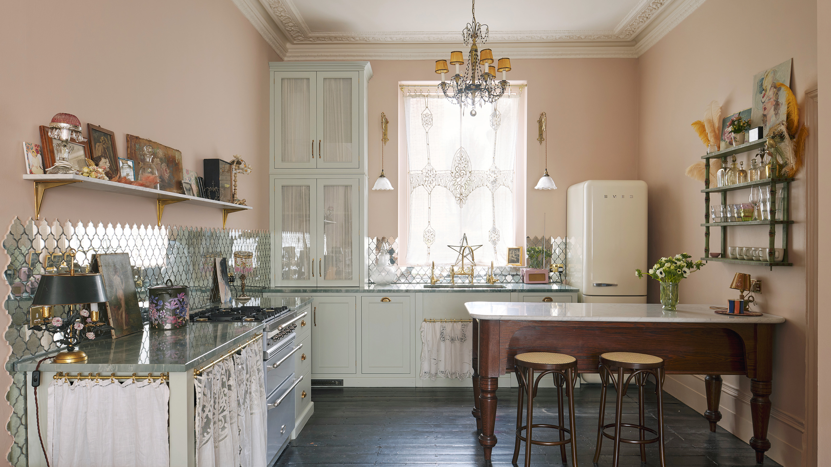 kitchen with pink walls and dark wood floors smeg fridge and lace covered window on the back wall with grey cabinetry below mirrored splashback on left wall wooden farmhouse table with stools and open shelving unit