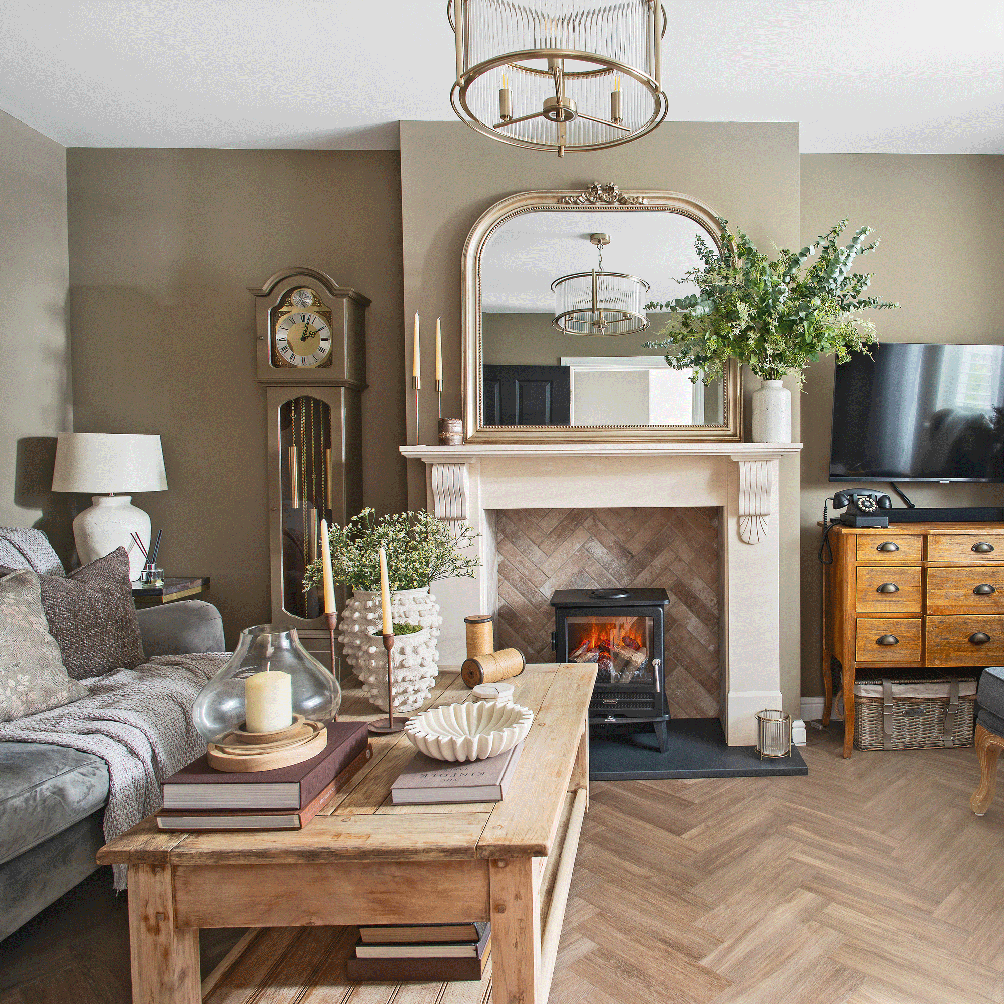 a neutral living room with herringbone wooden flooring a traditional fireplace with woodburner and an overmantel mirror beside a grandfather clock and a sofa