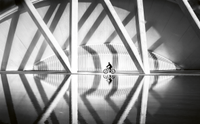 A cyclist rides along a waterside path beneath the angled white concrete ribs of a vast modern structure, with the whole scene mirrored in the still water below.
