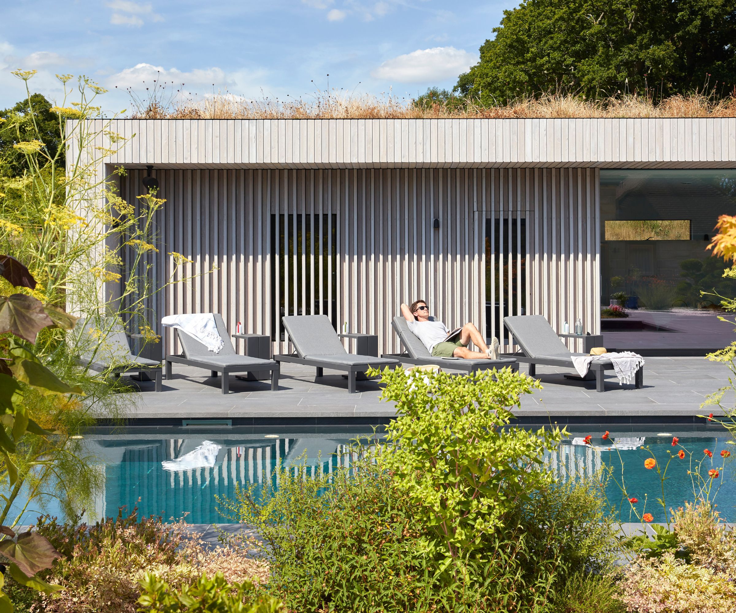 A man reading on a grey sun lounger beside a rectangular pool in front of a pale timber-clad pool house with a sedum green roof and large glazed doors.