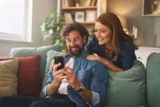 Couple shares a joyful moment in their new living room, smiling as they browse their smartphones, surrounded by comfortable decor and soft lighting.