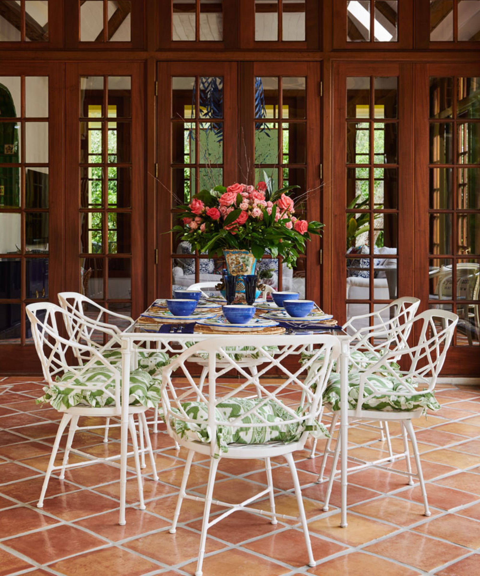 Outdoor patio with wooden glass doors leading out into the garden with a white metal dining table and matching chairs with green frilly cushions and a large rose arrangement at the centre of the table