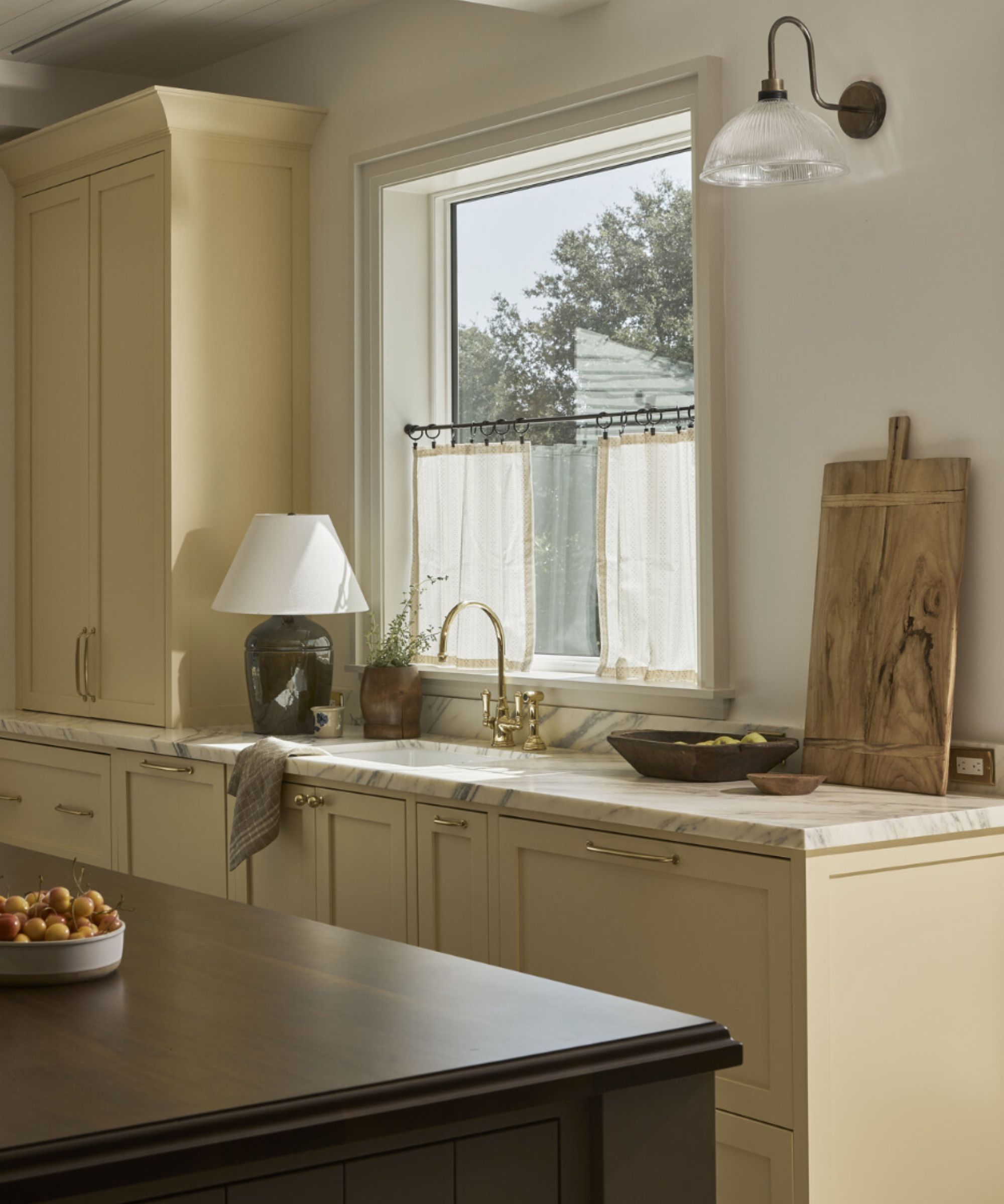 Kitchen with butter yellow cabinetry, marble countertop, dark wood island, counter decorated with lamp, chopping board and wooden bowls, window with cafe curtains above sink
