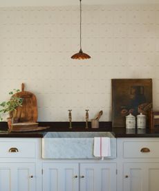 A kitchen with white lower cabinets, aged brass hardware, a black countertop, marble sink, and deVOL's new ditsy delft wallpaper in sepia on the wall