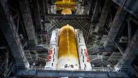 a large orange core stage of a booster is seen from below as it stands with two side mounted white solid rockets inside a high bay of an assembly building.