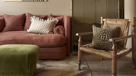 A living room with a pink sofa, green fabric footstool, and a woven arm chair with a leopard print cushion on it.