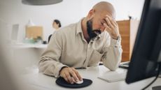 Man sitting at desk in front of computer rubs his forehead and looks tired