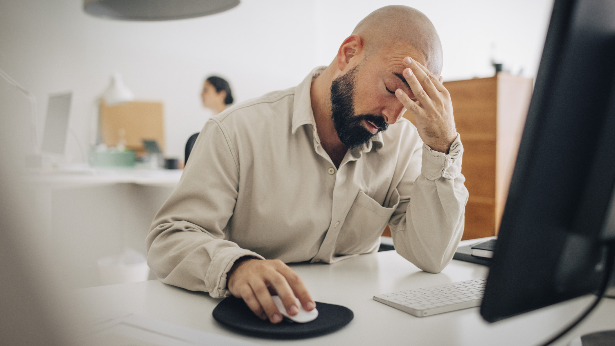 Man sitting at desk in front of computer rubs his forehead and looks tired