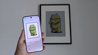 SwitchBot AI Art Frame hung on a plain white wall with a Pixel 10 Pro held up in front of it with the same Android bot artwork