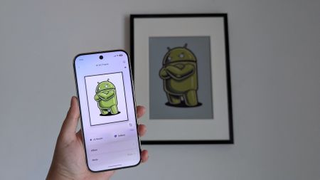 SwitchBot AI Art Frame hung on a plain white wall with a Pixel 10 Pro held up in front of it with the same Android bot artwork