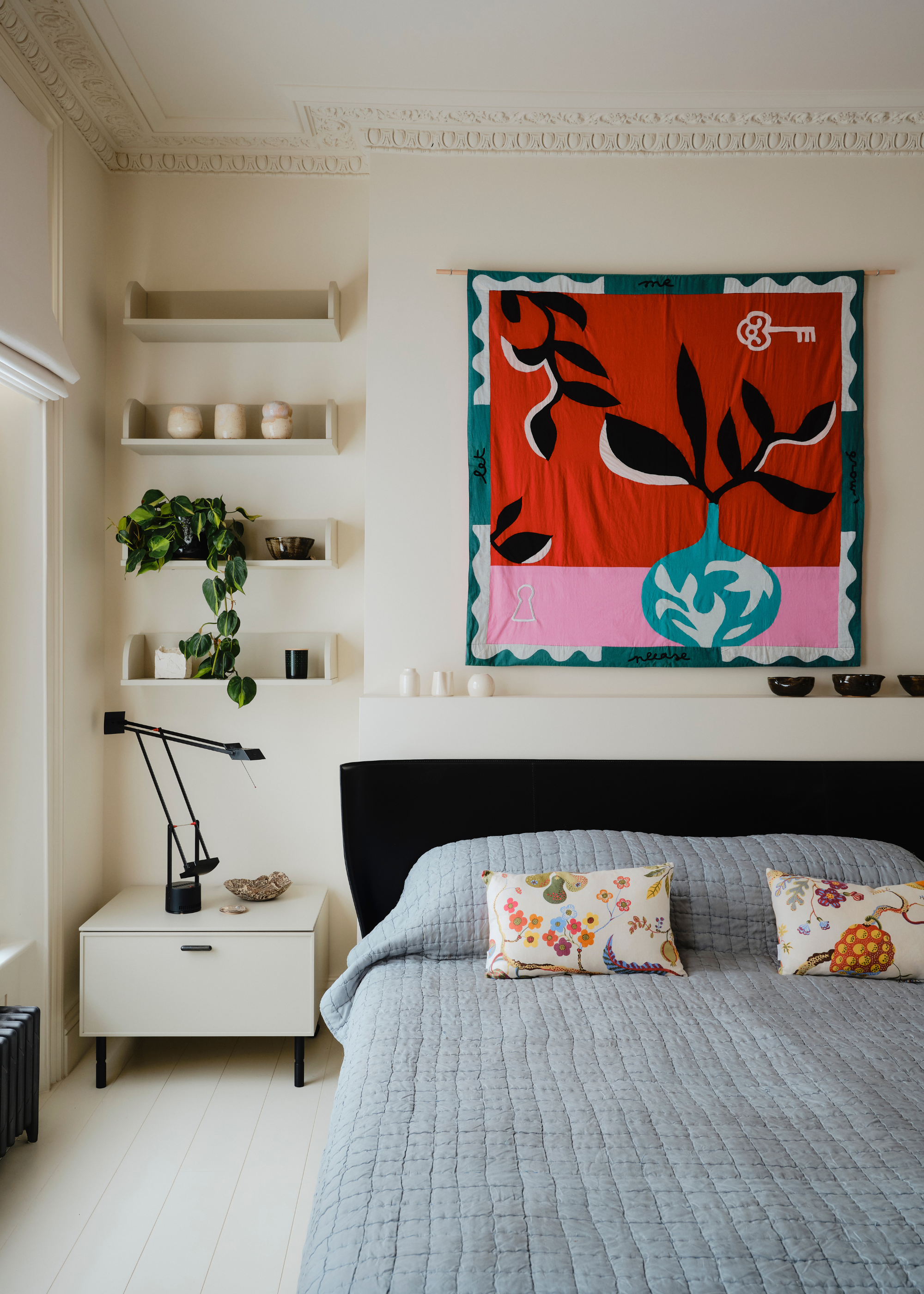 A white bedroom with floating shelves a minimalist side table and a large colorful tapestry above the blue bed