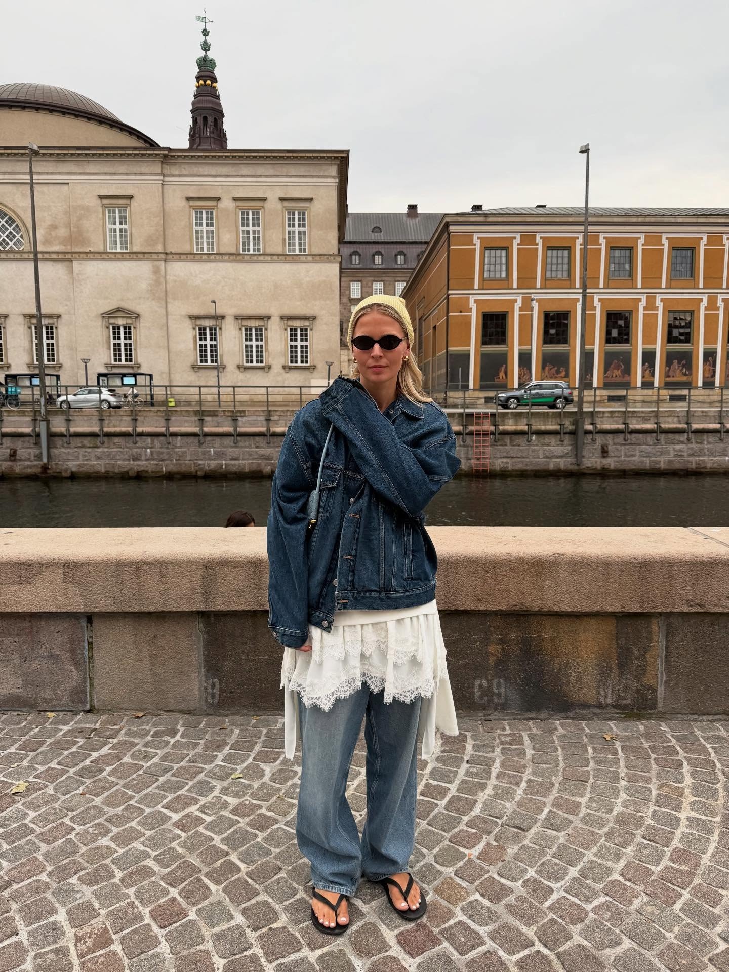 Influencer @holliemercedes wears a lace top over jeans and underneath a denim jacket. She accessories with black flip flops, a yellow headscarf and black sungalsses.