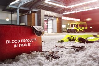 An image of the outside of a snow covered Holby ED at night with a bloody paramedic jacket lying on the ground beside a bag that reads "Blood products in transit."