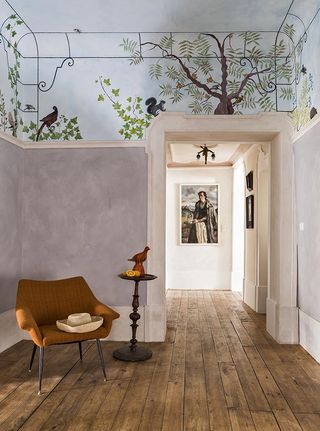 Interior view of a space at House of Birds featuring wooden floors and grey walls with a hand painted section at the top of a tree, greenery, birds and a squirrel against a blue background. There is also a dark orange chair with a hat on it, a dark stand with a bird statue and lemons and framed art on the walls