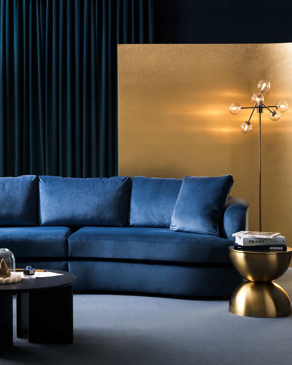 Image of a room that is navy blue with light blue carpeting. There is a shiny, gold room divider and a matching side table. There is a curved, cobalt blue sofa with a round, black coffee table in front of it.