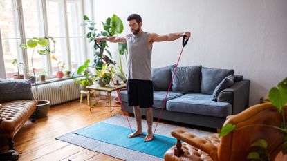 man in a living room setting on an exercise mat holding up resistance bands with handles laterally to shoulder height