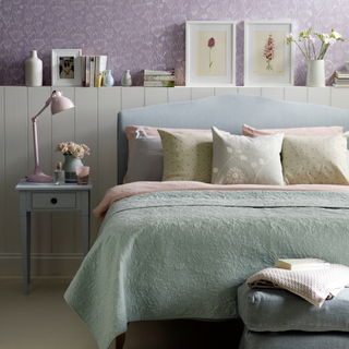 Pastel bedroom, vibrant purple wallpaper with floral pattern, decorative dispaly on top of tongue and groove panelled half wall, blue bedside tale, matching bedframe, assorted pastel bedlinen